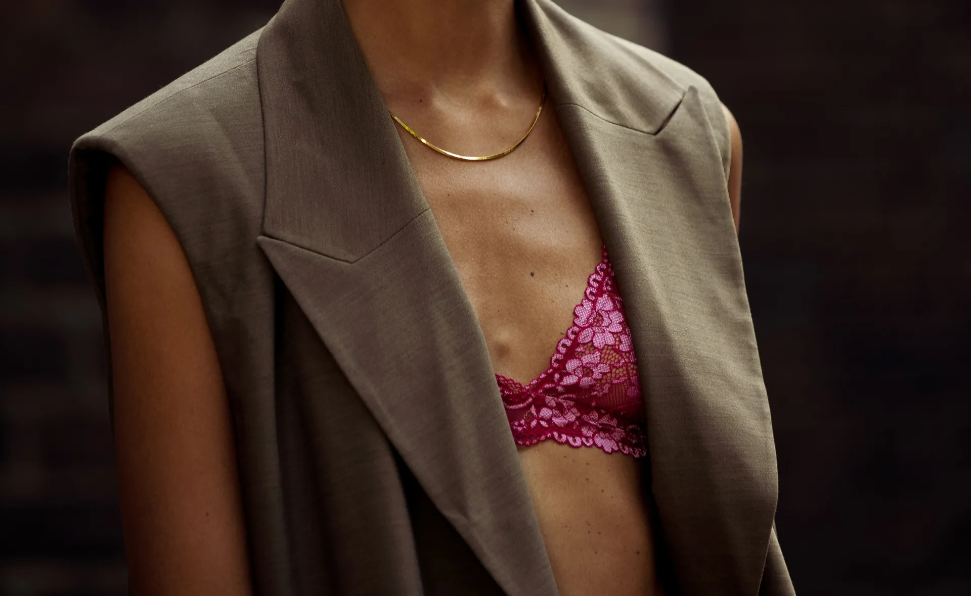 Woman in brown blazer with visible pink lace bra underneath, wearing a thin gold necklace.