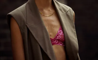 Woman in brown blazer with visible pink lace bra underneath, wearing a thin gold necklace.