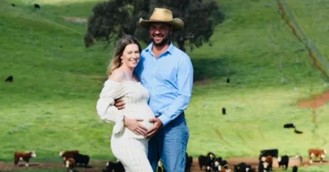 Pregnant couple smiling, standing in a green pasture with cows in the background. Man in cowboy hat embraces woman gently.