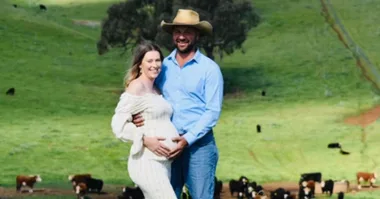 Pregnant couple smiling, standing in a green pasture with cows in the background. Man in cowboy hat embraces woman gently.