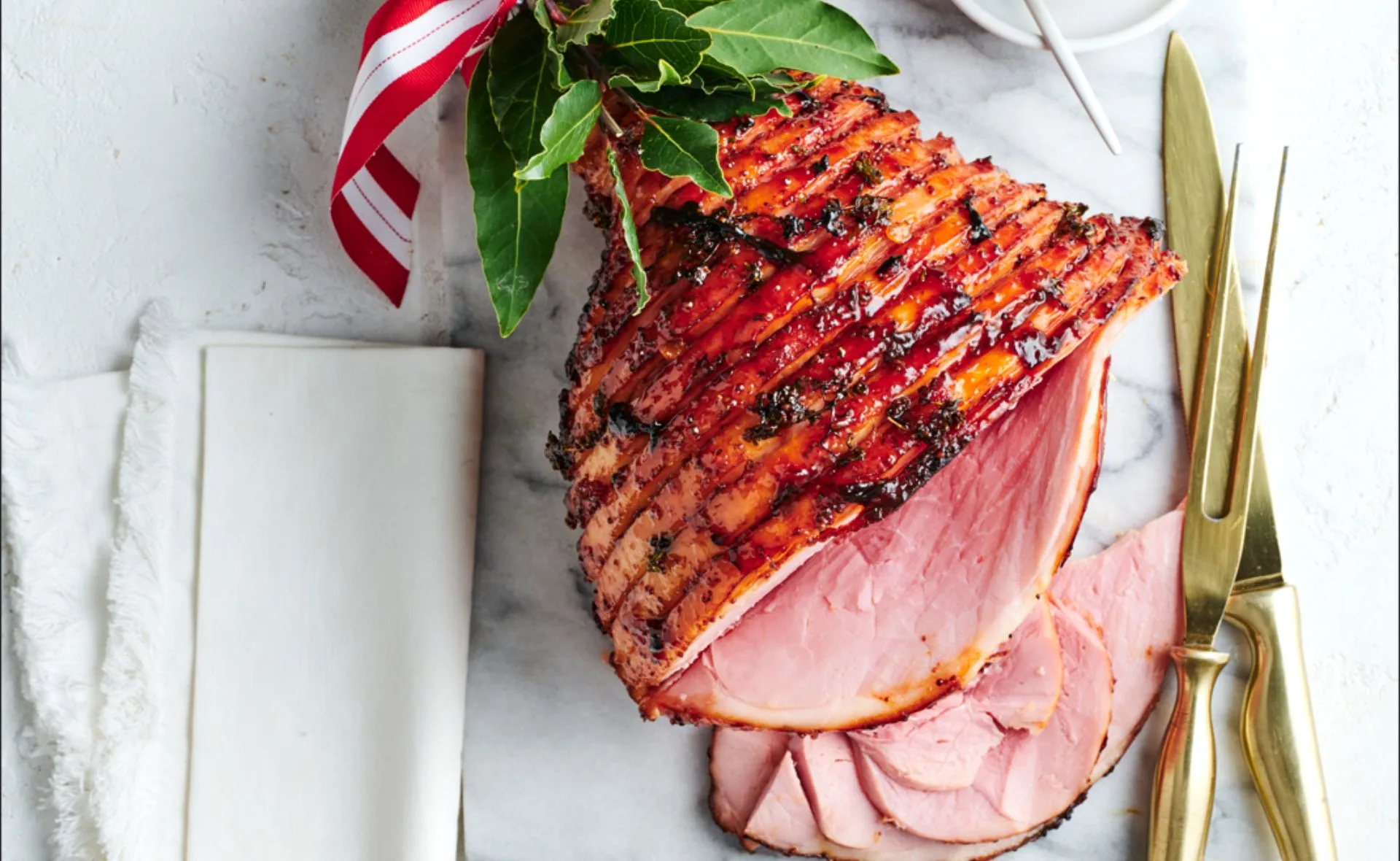 Glazed Christmas ham with a red ribbon, garnished with herbs, on a marble board next to gold utensils.