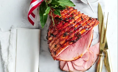 Glazed Christmas ham with a red ribbon, garnished with herbs, on a marble board next to gold utensils.