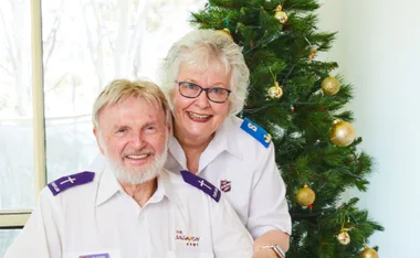 Elderly couple in Salvation Army uniforms smiling by a decorated Christmas tree indoors.