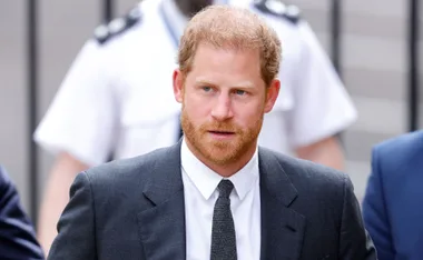 Prince Harry wearing a suit and tie, walking outdoors with blurred figures in the background.