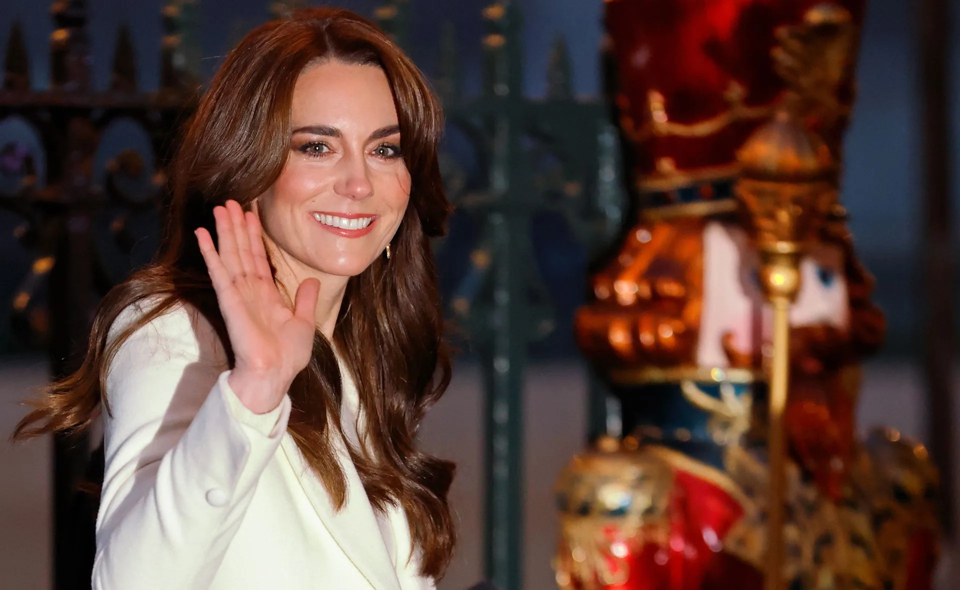 Person in a white coat smiling and waving, with festive decorations in the background.