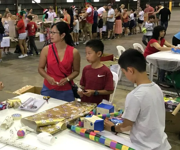 People wrapping gifts at a charity event, with tables full of wrapping paper and supplies in a large indoor space.
