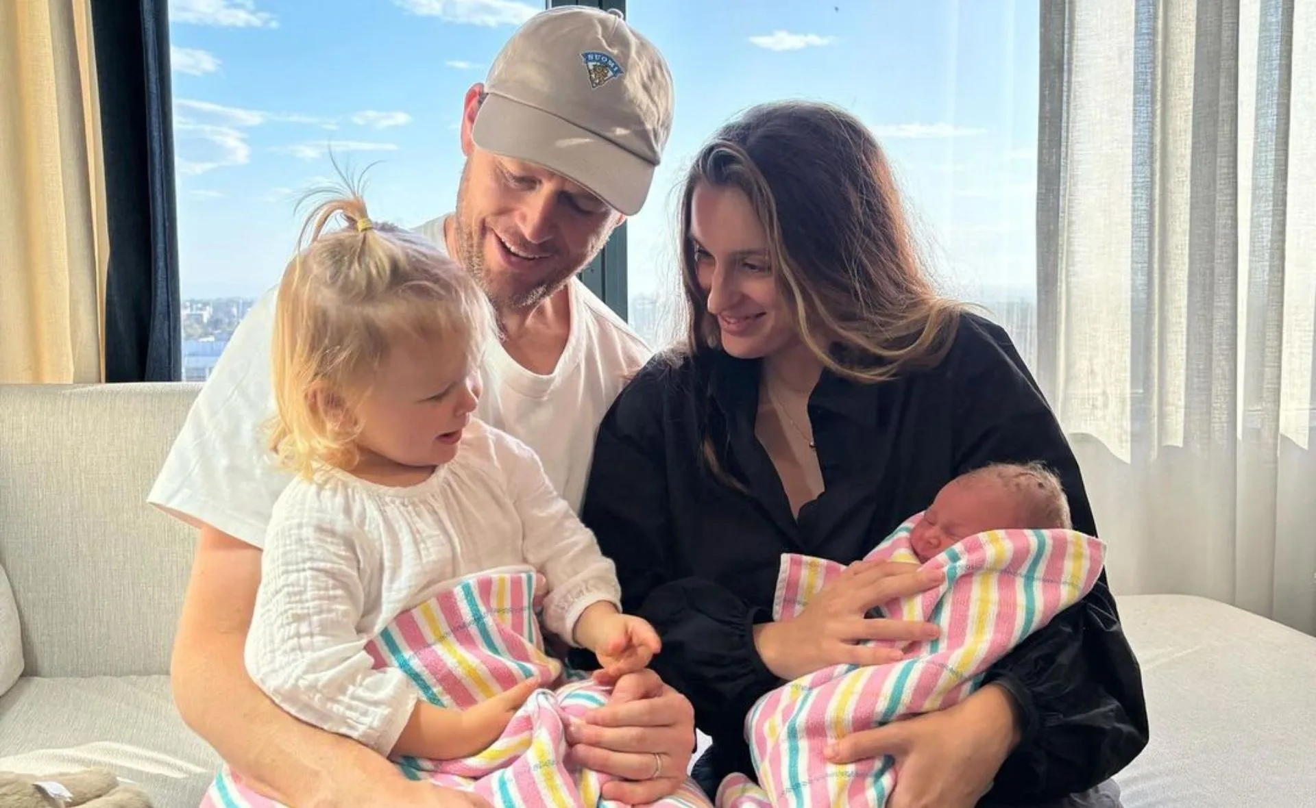 A family of four sits on a couch by a window, smiling and holding two children wrapped in colorful blankets.