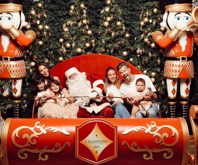 Family posing with Santa Claus on a sleigh, flanked by nutcracker figures, Christmas trees in the background.