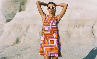 Woman in a geometric patterned dress and sunglasses standing in a sandy landscape with arms behind head.