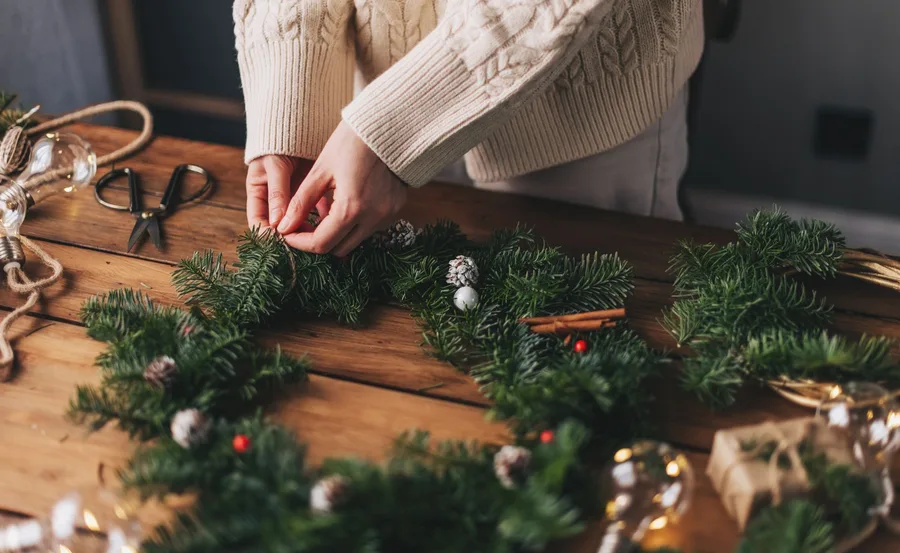 Person crafting a Christmas wreath with pine branches, cinnamon sticks, and small decorations on a wooden table.
