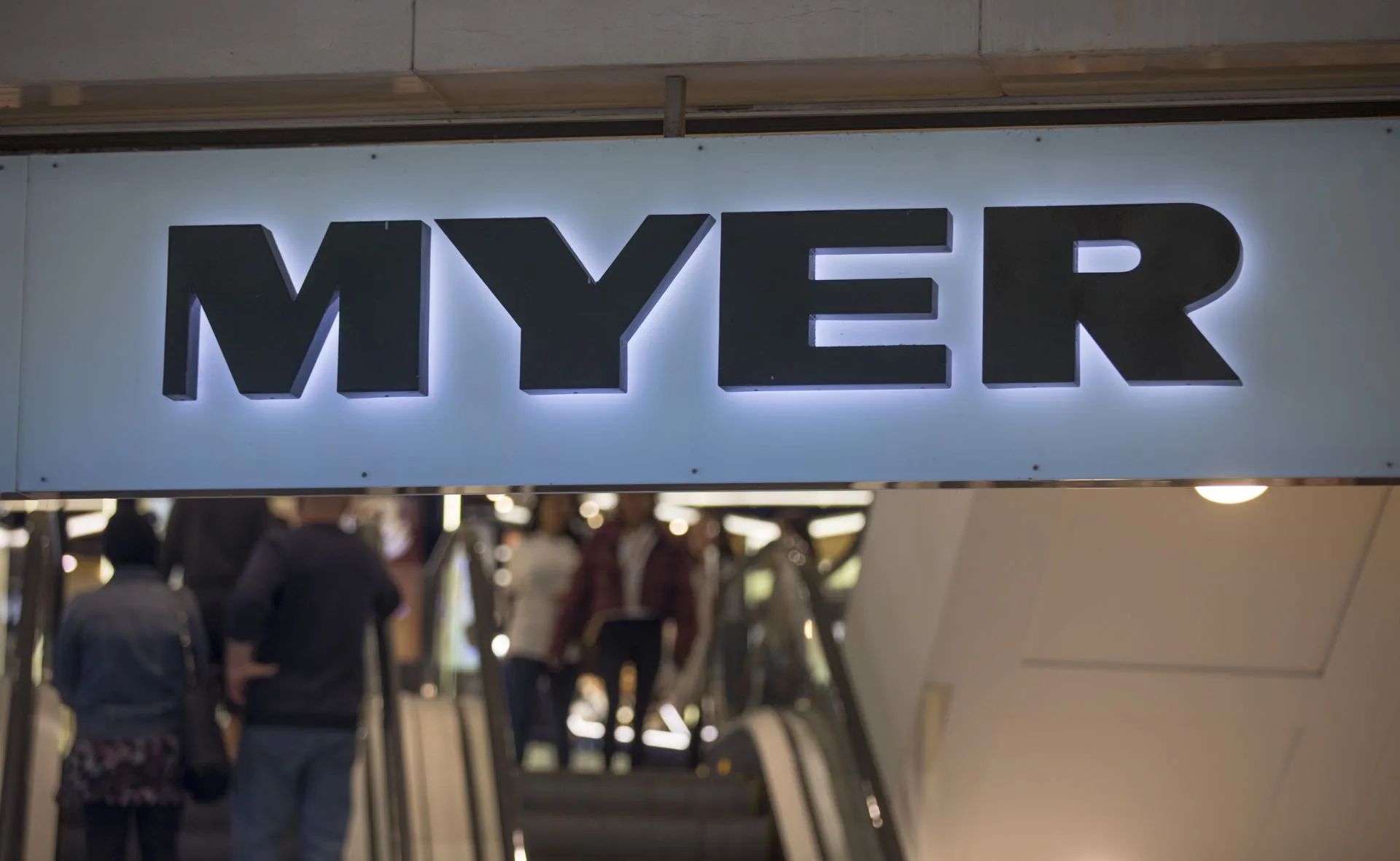 Sign for Myer department store above a busy escalator with shoppers.