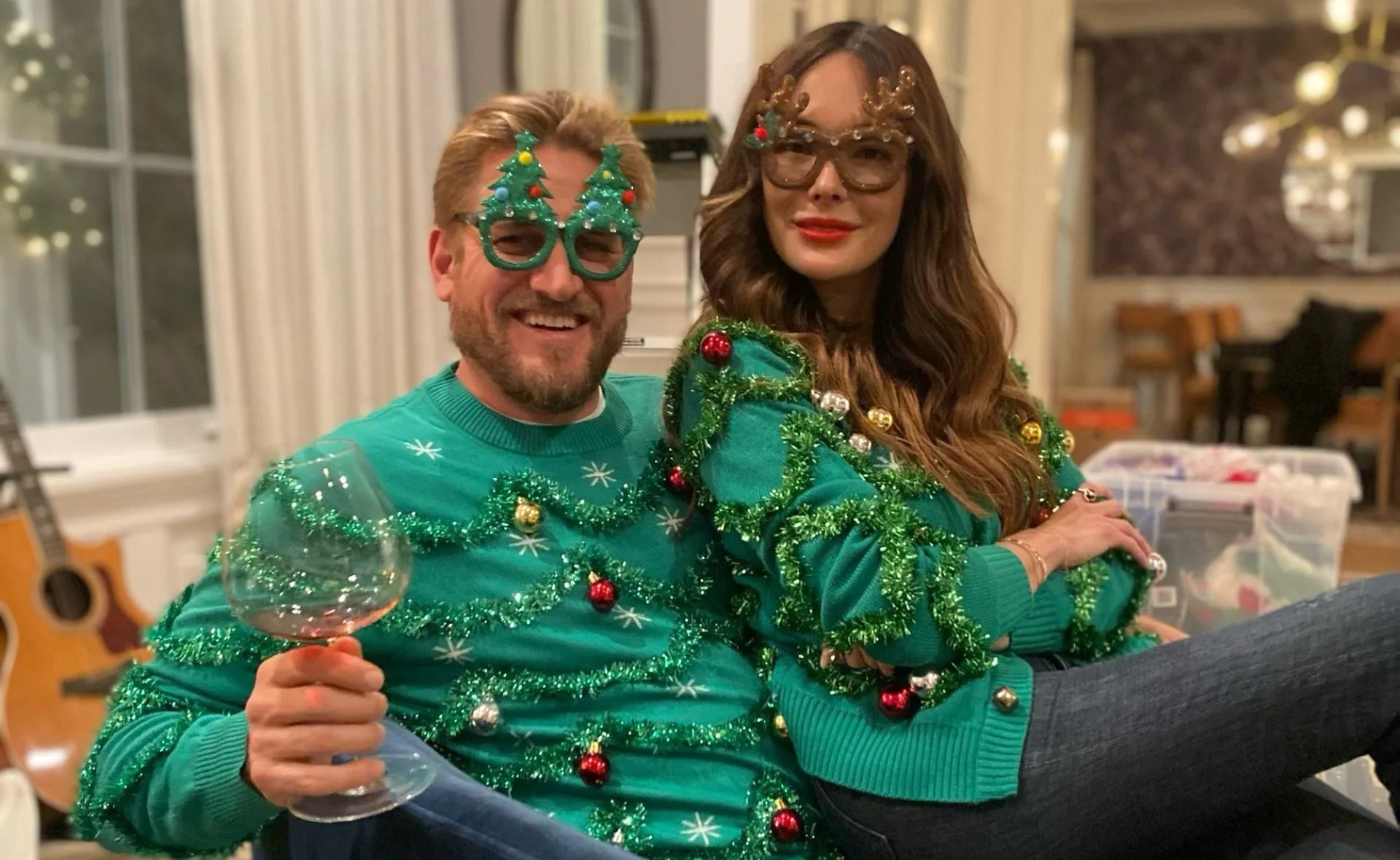 Couple wearing green Christmas sweaters and festive glasses, sitting together and smiling with a wine glass.