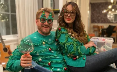 Couple wearing green Christmas sweaters and festive glasses, sitting together and smiling with a wine glass.