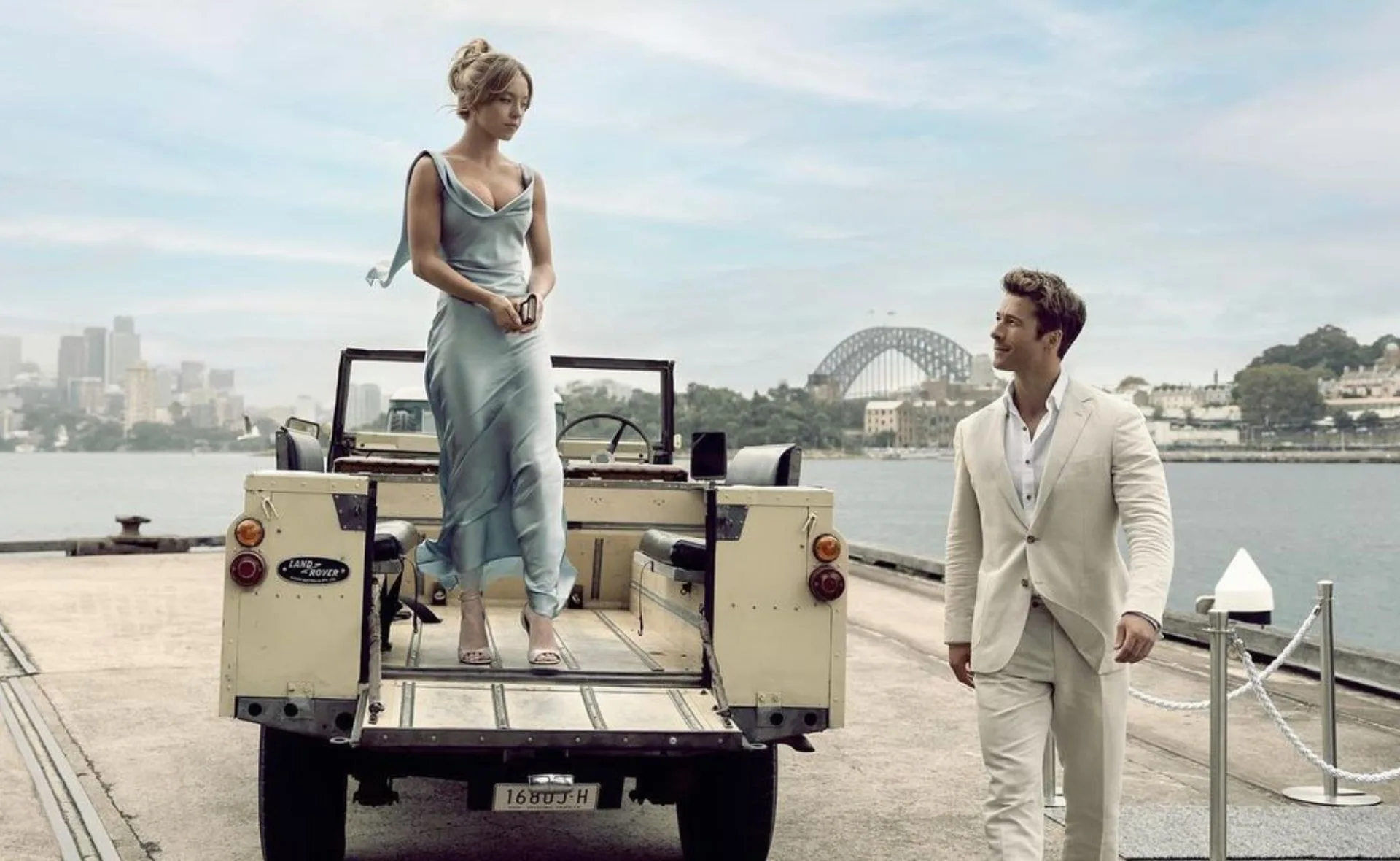 "Two people in elegant attire stand by a vintage Land Rover near Sydney Harbor."