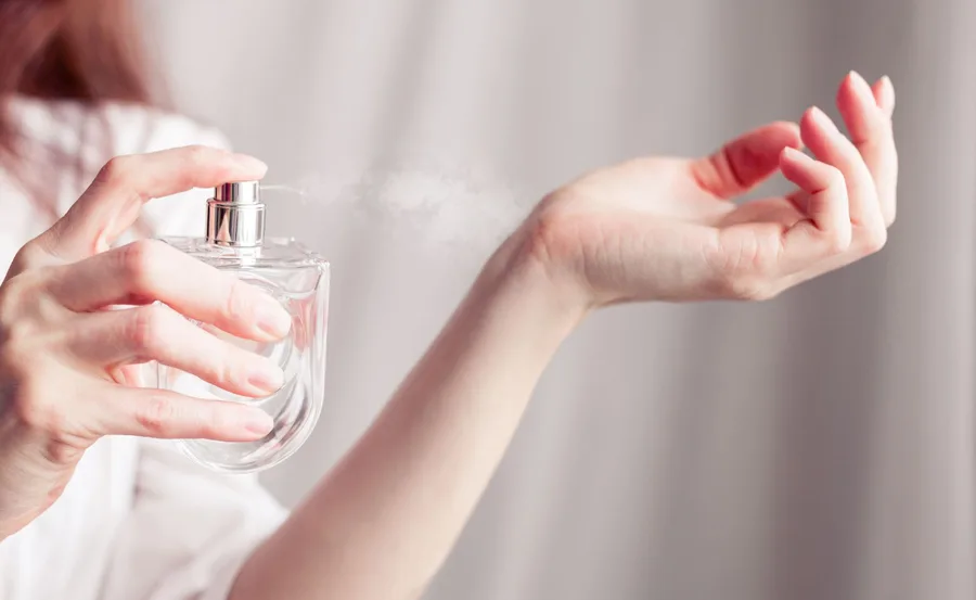 A person spritzing perfume onto their wrist from a transparent glass bottle, with a mist visible in the air.