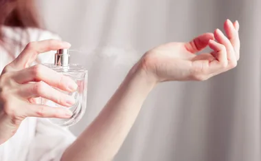 A person spritzing perfume onto their wrist from a transparent glass bottle, with a mist visible in the air.