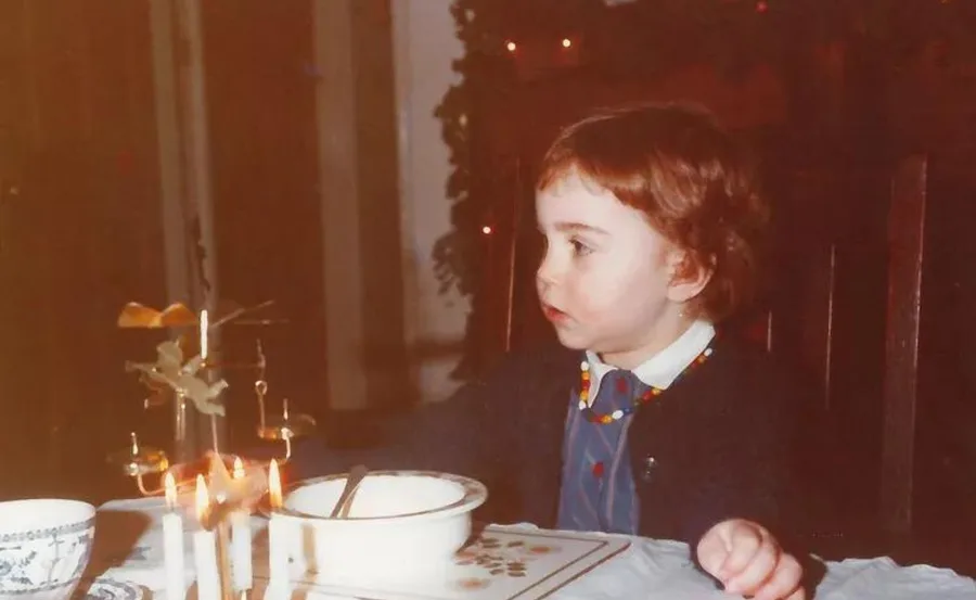 Young child sitting at a table with candles and a bowl, wearing a colorful necklace and looking to the side.