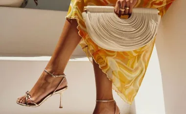 Woman in a yellow dress with gold heels, holding a white woven clutch on steps.