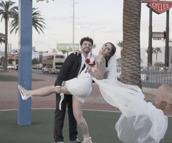 A couple in wedding attire poses playfully on a sunny street, with palm trees and signs in the background.