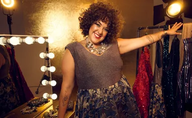 Woman in a sparkly gown smiles near a vanity mirror, with elegant dresses on a rack in the background.
