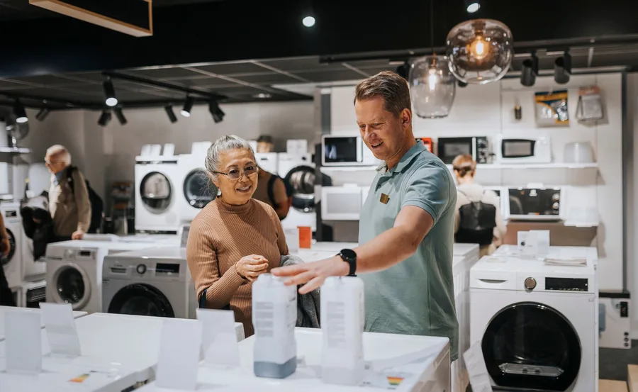Salesperson assisting a customer while shopping for appliances in a store, surrounded by washing machines.