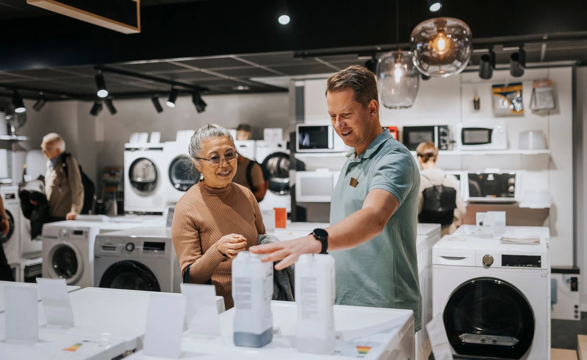 Salesperson assisting a customer while shopping for appliances in a store, surrounded by washing machines.