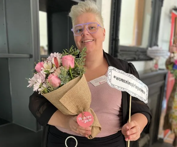 Person with short hair holds a bouquet of pink flowers and a sign reading "#womeninspiringwomen."