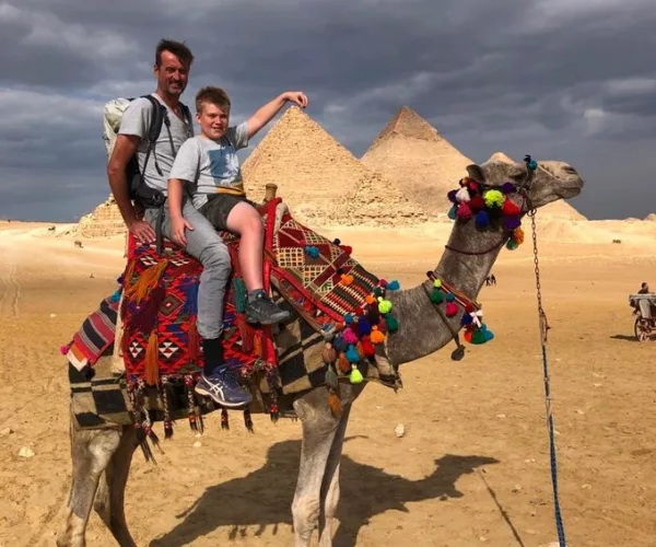 Father and son riding a camel with Pyramids of Giza in the background on a sunny day.