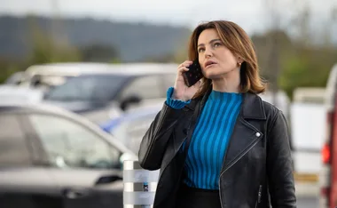 Woman in a black leather jacket talking on a smartphone outdoors, with blurred cars in the background.