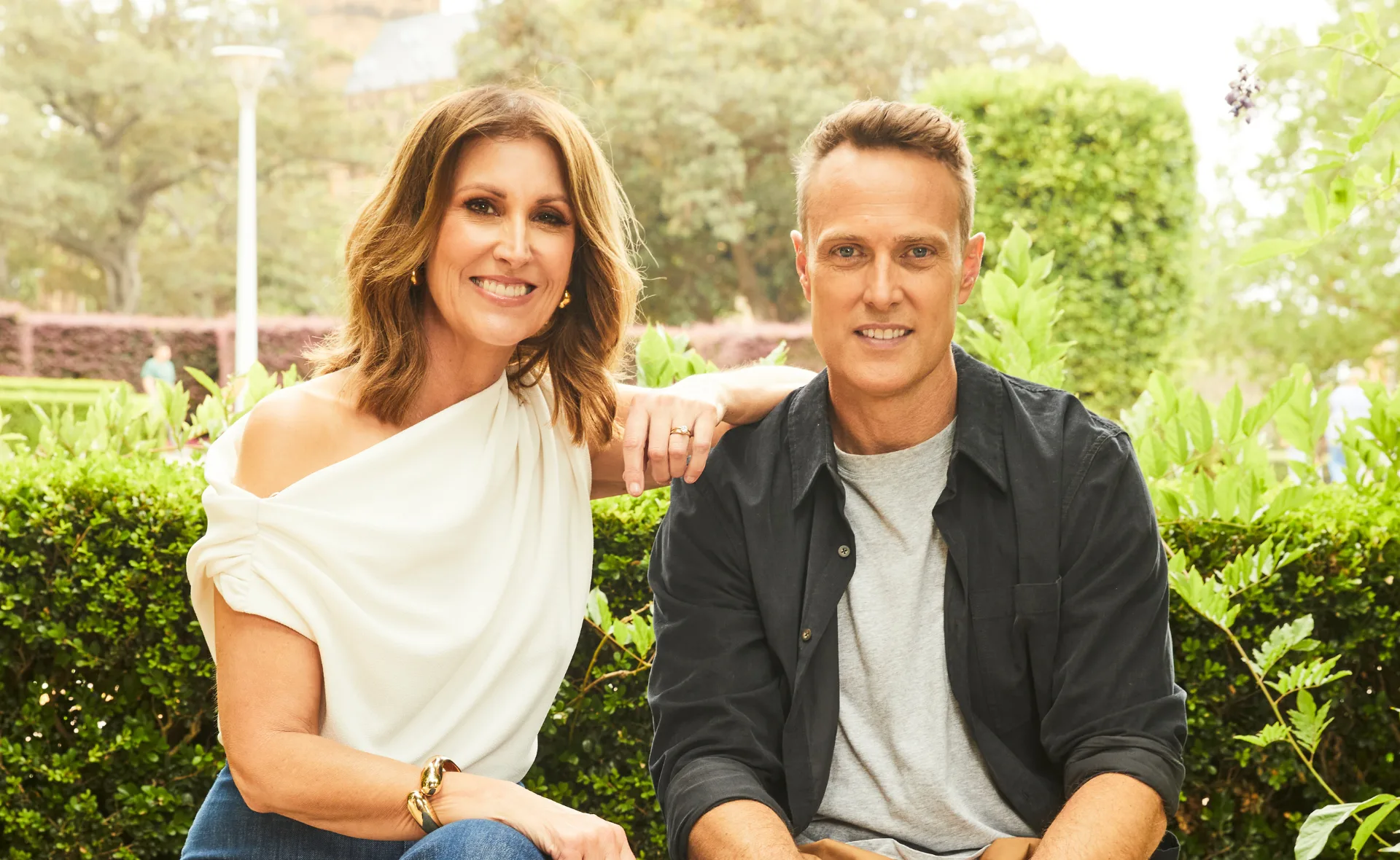 Two people smiling seated outdoors, with greenery in the background.