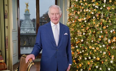 A person in a blue suit stands by a decorated Christmas tree inside a room with a large window.