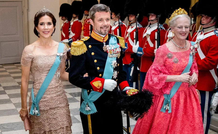 Royal figures pose in formal attire with sashes and crowns, flanked by guards in uniform at a ceremonial event.