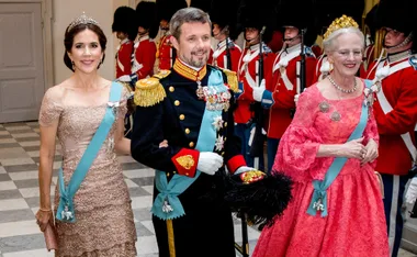 Royal figures pose in formal attire with sashes and crowns, flanked by guards in uniform at a ceremonial event.