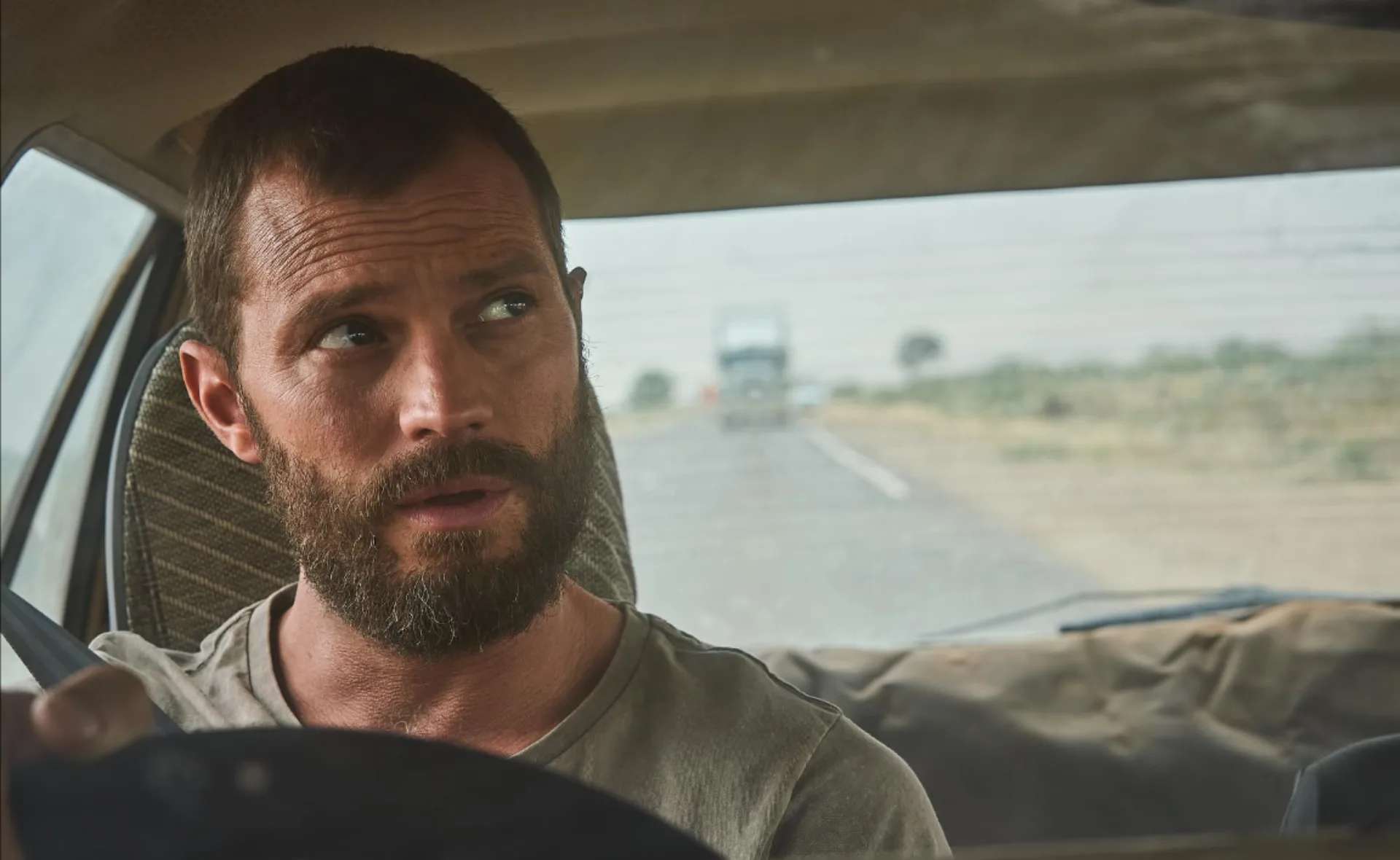 A bearded man with short hair sits behind the wheel of a car, looking to his left with a serious expression.