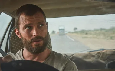 A bearded man with short hair sits behind the wheel of a car, looking to his left with a serious expression.