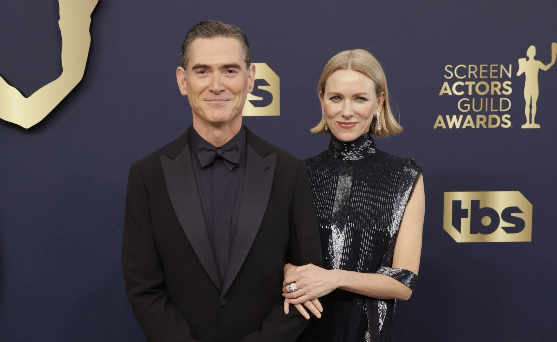 Man and woman posing in formal attire at the Screen Actors Guild Awards with dark background and logos.