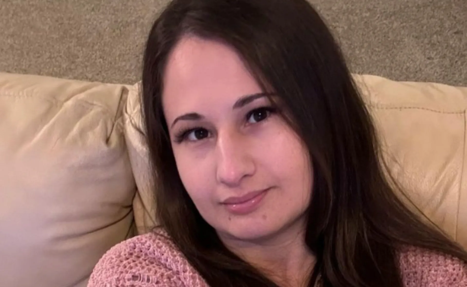Young woman with long brown hair sitting on a beige couch, wearing a pink sweater, looking towards the camera.
