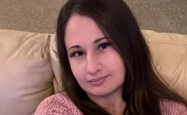 Young woman with long brown hair sitting on a beige couch, wearing a pink sweater, looking towards the camera.