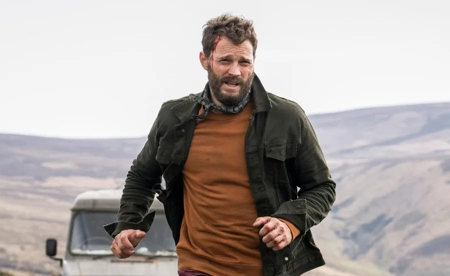 Man running with a rugged appearance in a rural setting, wearing an olive jacket and orange shirt.
