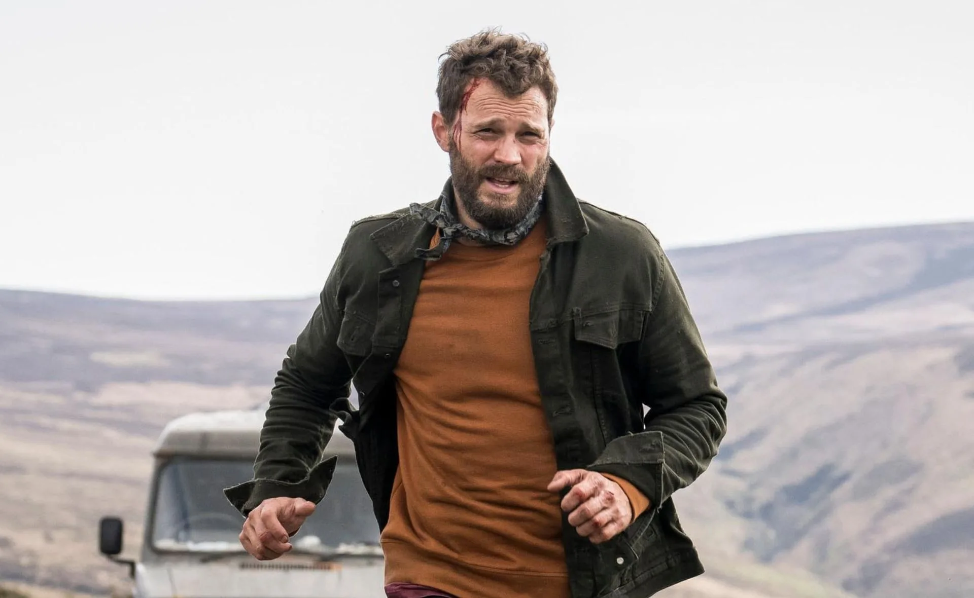 Man running with a rugged appearance in a rural setting, wearing an olive jacket and orange shirt.