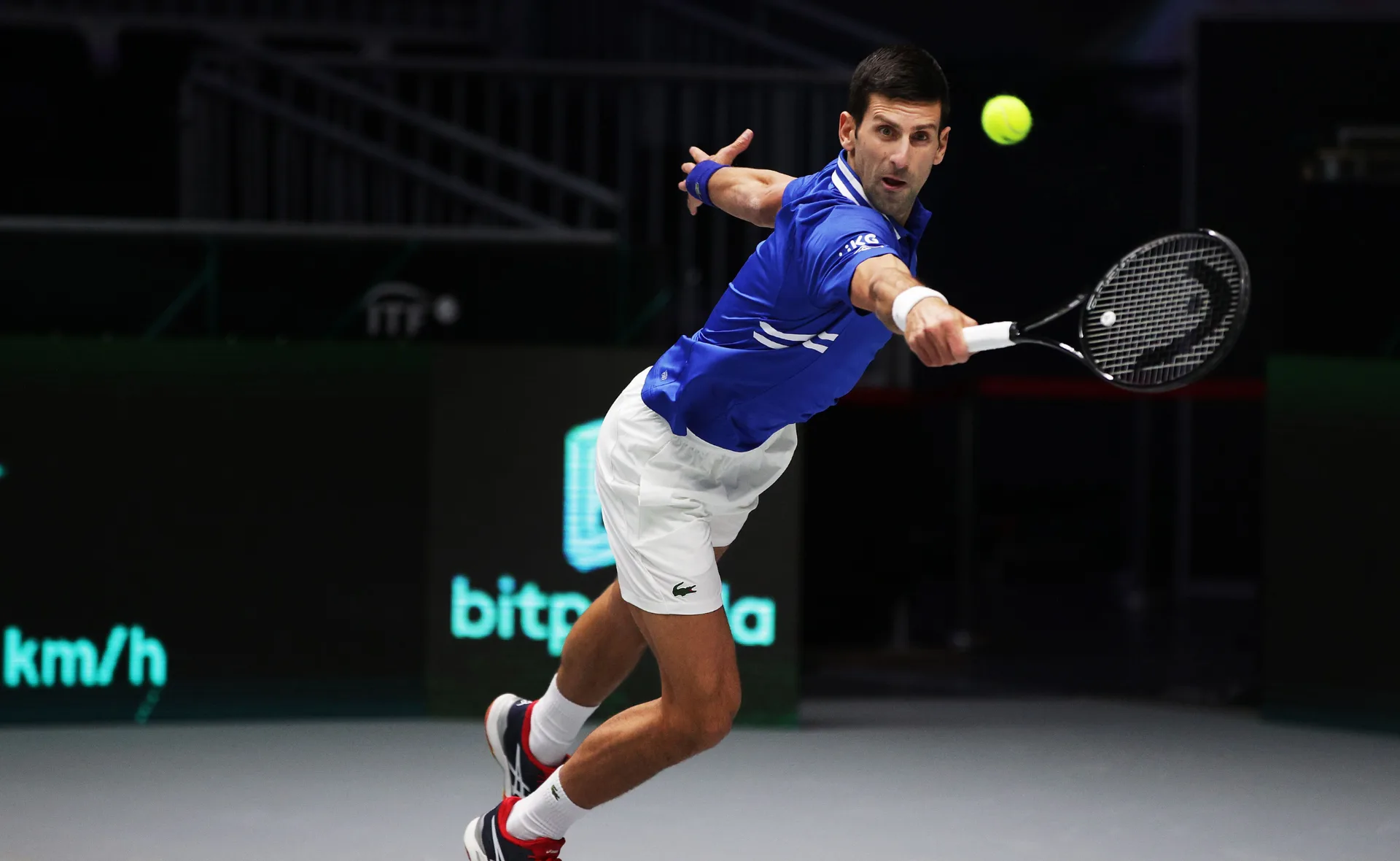 Tennis player in blue shirt lunges to hit a forehand during a match on an indoor court.