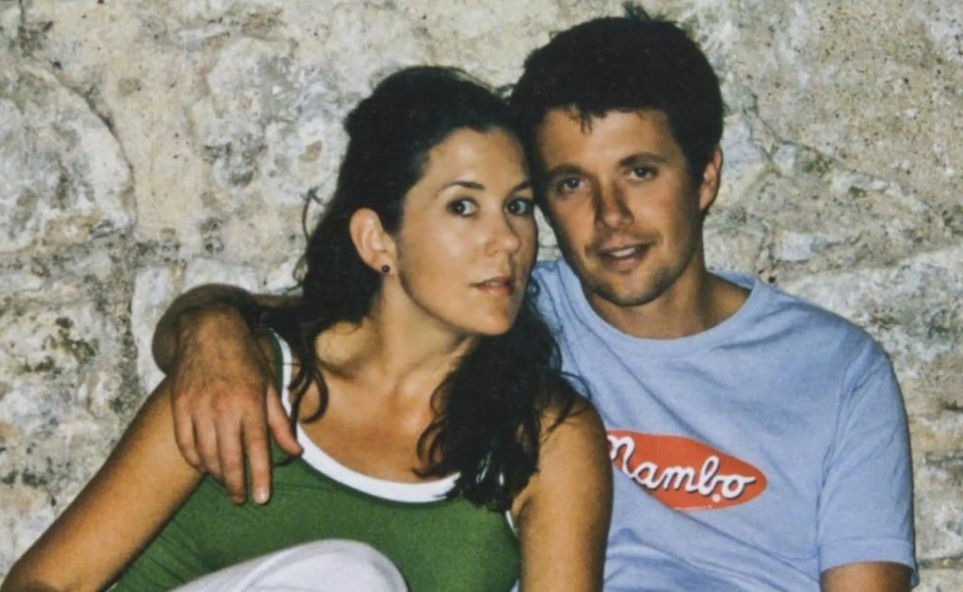 A man and woman sitting close, smiling against a textured stone wall.