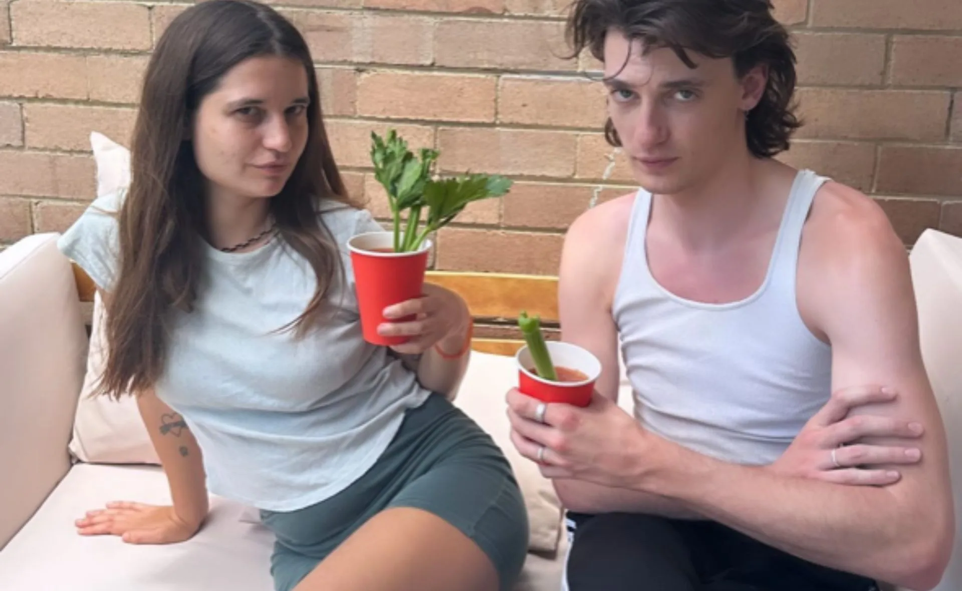 Two people sitting on a couch, holding red cups with celery, posing casually against a brick wall.