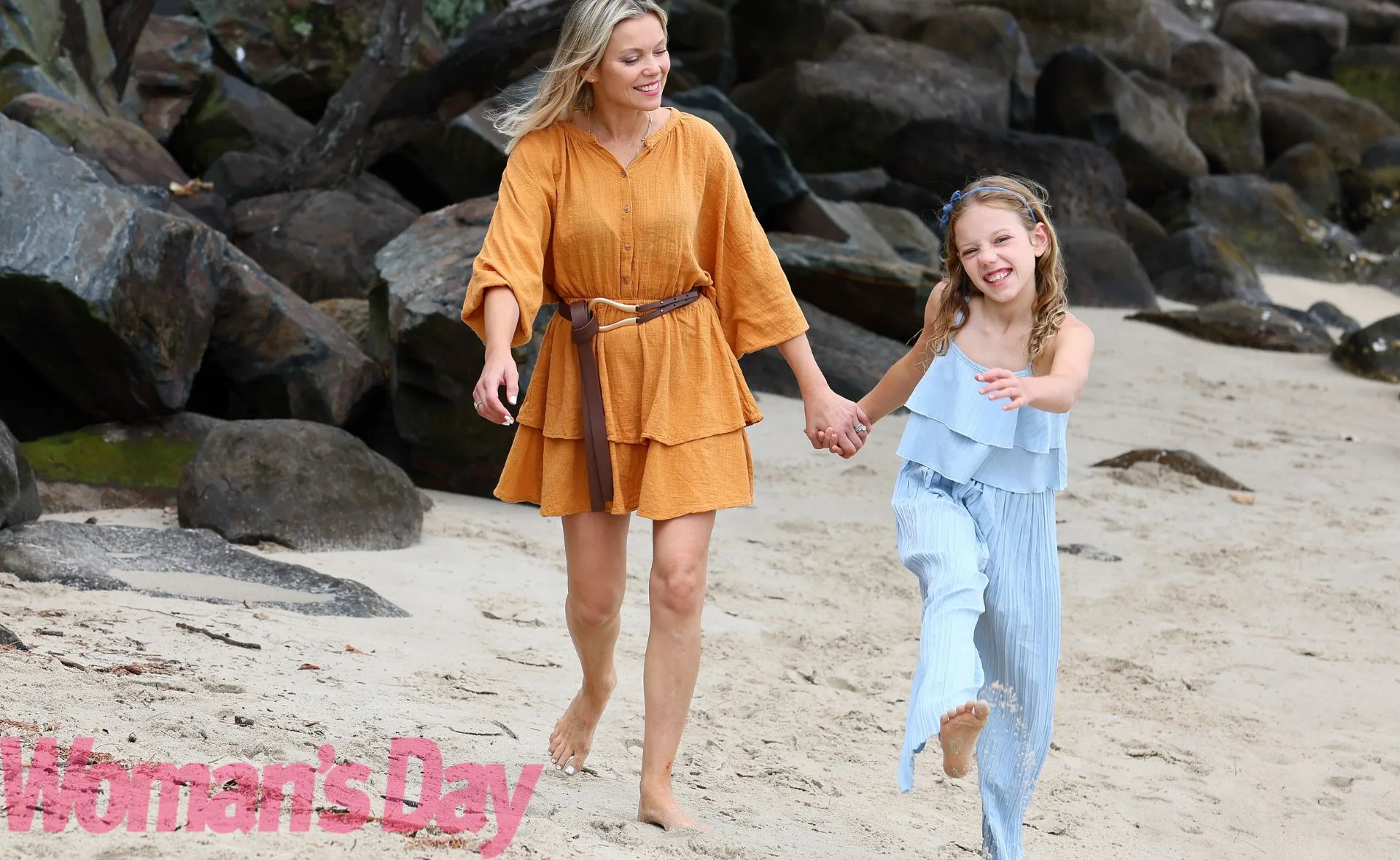 Mother and daughter walk joyfully on the beach, holding hands and smiling, surrounded by rocks.