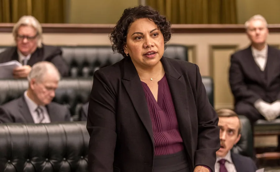 A woman in a purple blouse and black suit speaks confidently in a parliamentary setting, surrounded by seated individuals.