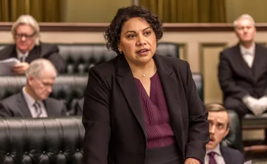 A woman in a purple blouse and black suit speaks confidently in a parliamentary setting, surrounded by seated individuals.
