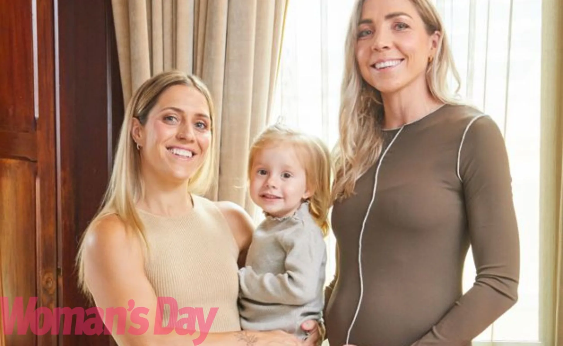 Three women smiling, one holding a young child, in an indoor setting with curtains in the background.