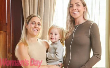 Three women smiling, one holding a young child, in an indoor setting with curtains in the background.