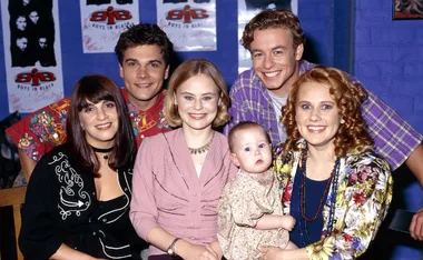 Group of six people, including a baby, smiling indoors with "Boys in Black" posters on the wall behind them.