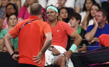 Man in red shirt sitting with a pained expression, holding his side, while another person in orange speaks to him.